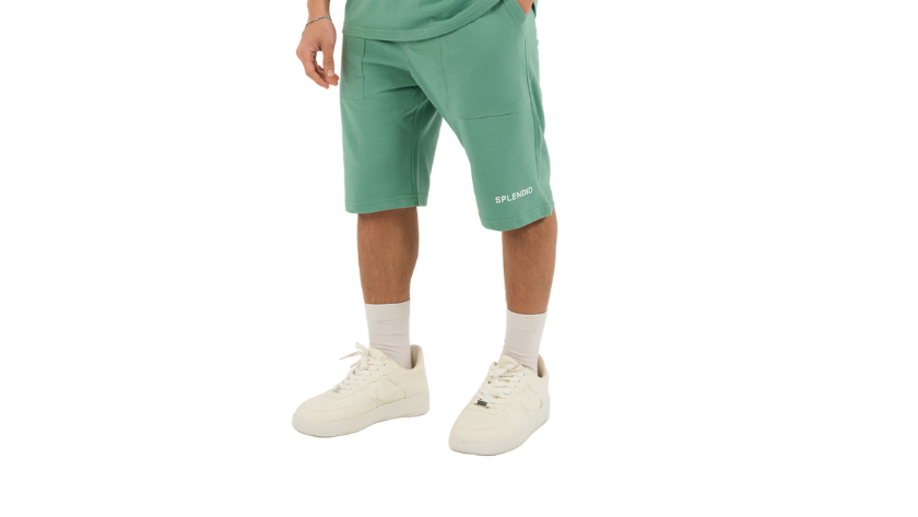 A male model wearing a matching green athletic set consisting of a short-sleeve polo shirt and matching shorts, paired with white socks and sneakers, against a plain white background.