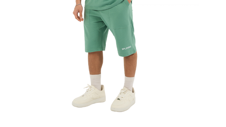 A male model wearing a matching green athletic set consisting of a short-sleeve polo shirt and matching shorts, paired with white socks and sneakers, against a plain white background.